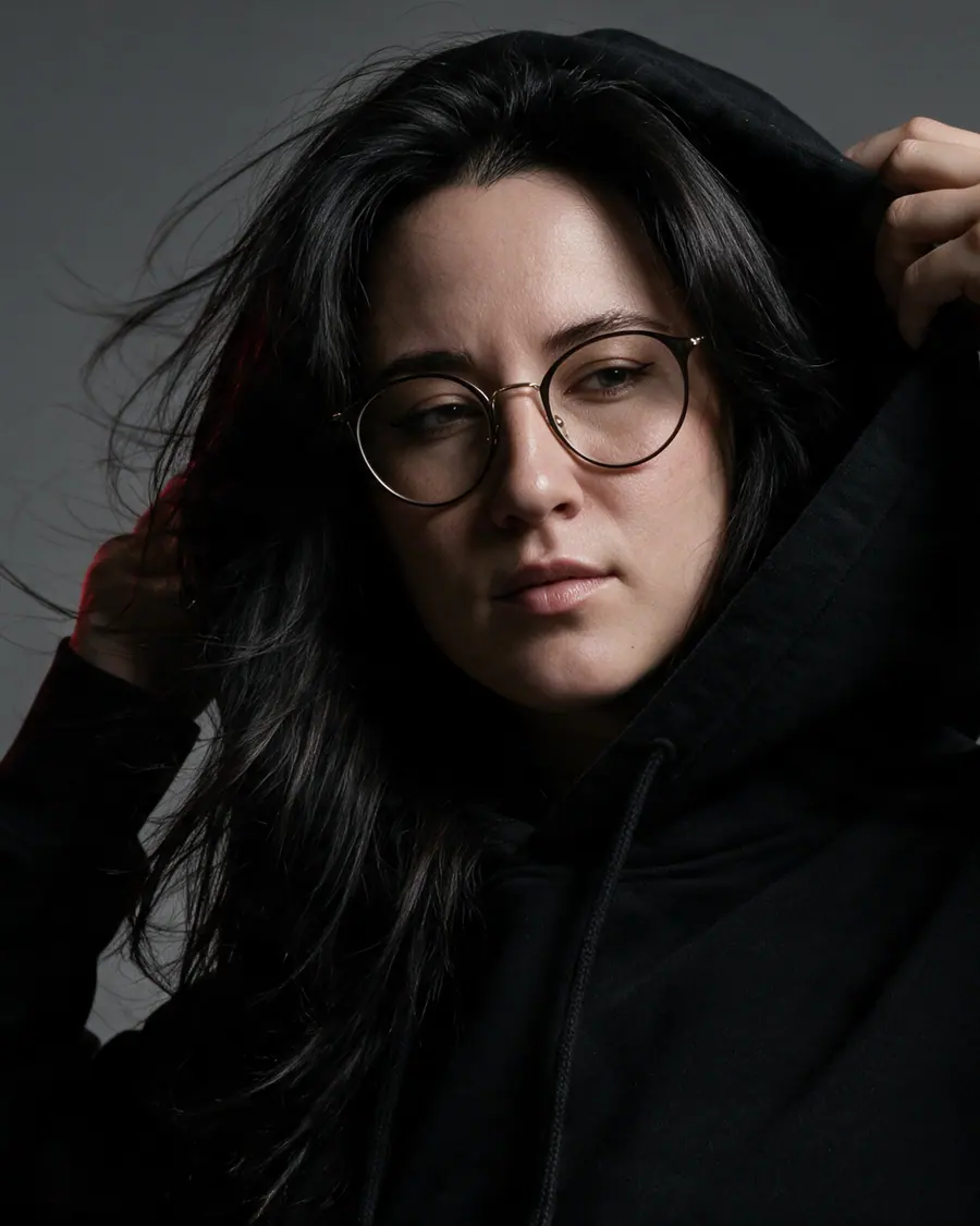 Catherine adjusting the hood of an oversized black garment in a moody editorial portrait.